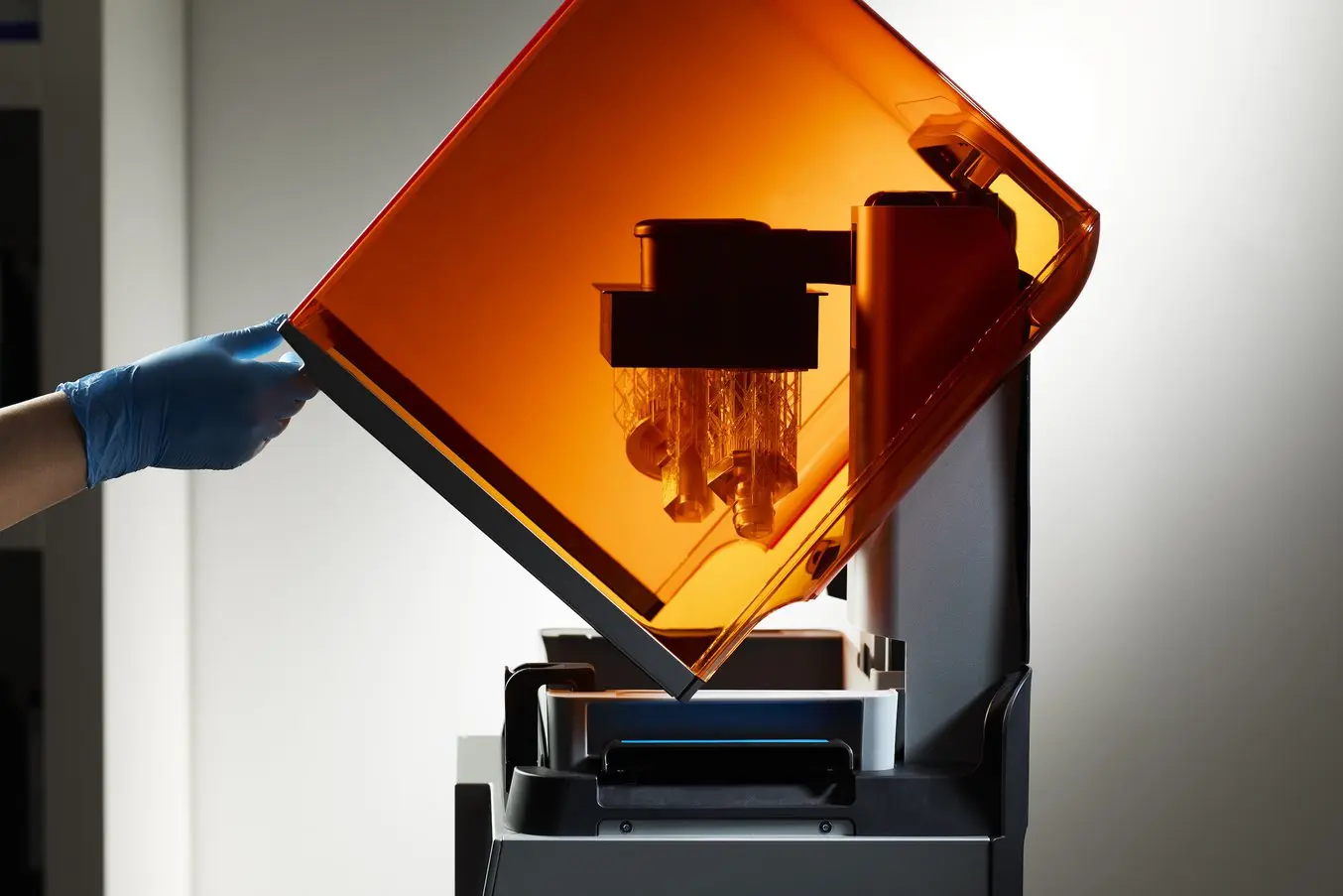 Una mano coperta da un guanto blu solleva il coperchio protettivo arancione di una stampante 3D Form 4